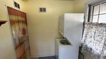 A compact kitchen with a washing machine, a small refrigerator, and a window with floral curtains.