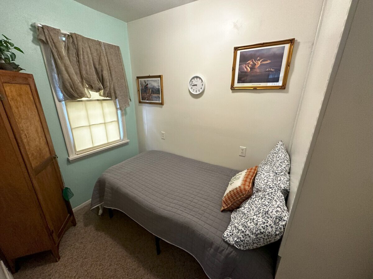 Un dormitorio con cama, ventana con cortinas, un reloj de pared y cuadros enmarcados.