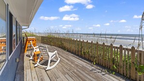 A wooden deck with a view of the beach, a wooden fence, and a white rocking chair. - Oceanfront Oasis w BBQ Pet Friendly Beach Access (Saint Augusitne)