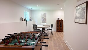 A room with a foosball table, a dining table with chairs, a cabinet, and a framed picture on the wall.