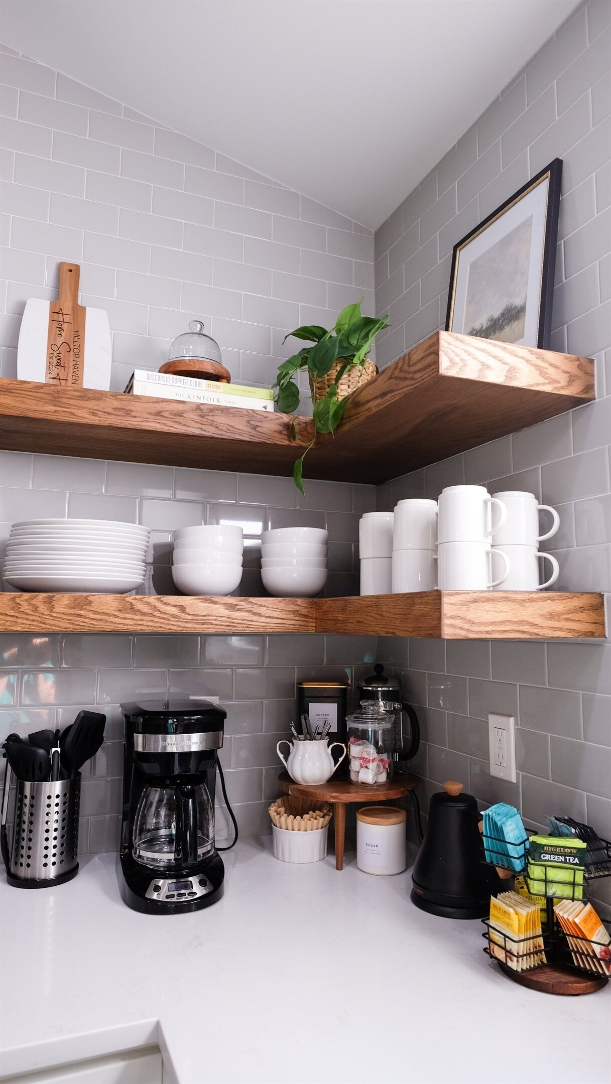 A kitchen with grey tiled walls, wooden shelves holding plates, bowls, and mugs, a coffee maker, and a plant.