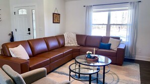 A living room with a brown leather sectional sofa, a glass coffee table, and a beige armchair.