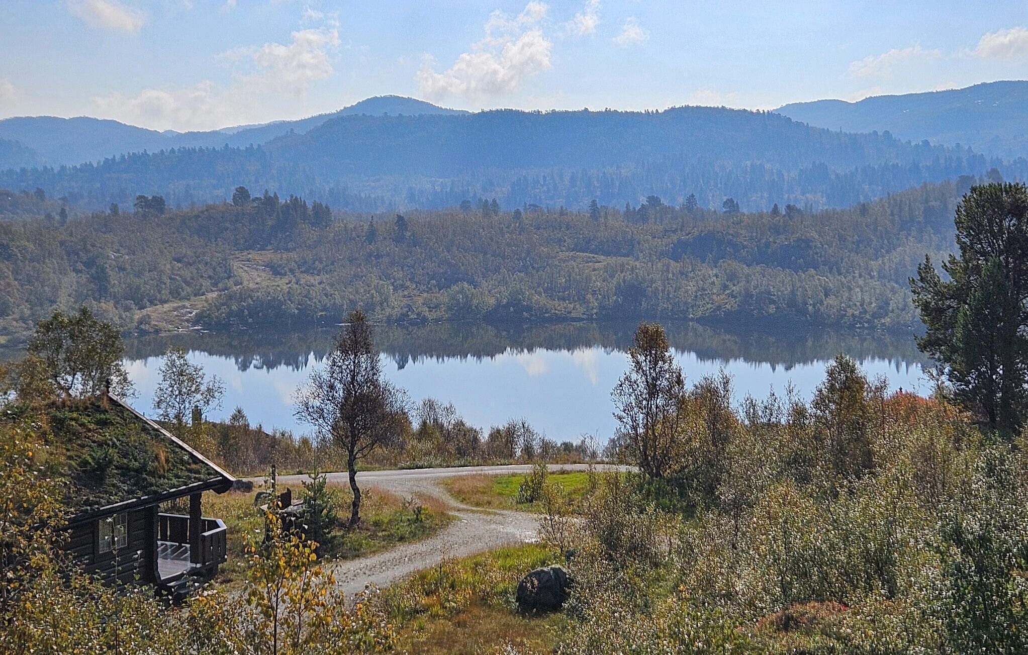 Una cabaña con porche, rodeada por un bosque y un lago, con montañas al fondo.