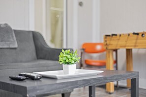 Un salon avec un canapé gris, une petite table blanche ornée d’un pot de plantes, et quelques télécommandes posées sur la table.