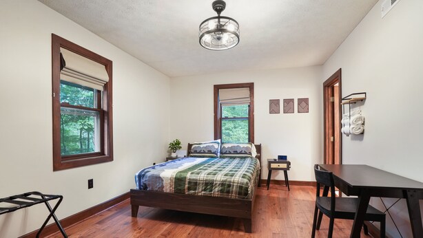 A bedroom with a bed, a desk, and a chair. There are windows with curtains, a ceiling fan, and a wall-mounted shelf with towels.