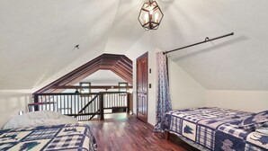 Quarto com duas camas, soalho de madeira, teto inclinado, escada e um candeeiro suspenso.