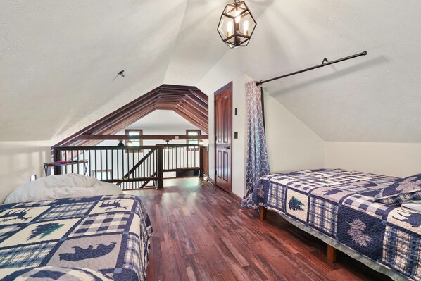 A bedroom with two beds, wooden flooring, a sloped ceiling, a staircase, and a hanging light fixture.