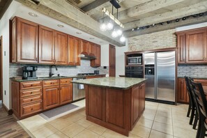 A kitchen with wooden cabinets, a granite countertop, a stainless steel refrigerator, and a brick wall.
