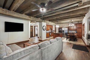 A spacious living room with a large gray sectional sofa, a flat-screen TV mounted on the wall, and a ceiling fan. The kitchen area is visible with wooden cabinets and a dining table.
