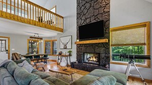 A spacious living room with a large sectional sofa, a stone fireplace, a mounted TV, and a balcony with a telescope. - Moose Shack - Mountain Views · Spa King Suite · Game Room · 3 Patios (Blairsville)