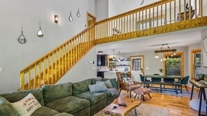 A spacious living area with a green sofa, a wooden staircase, a dining table, and a kitchen in the background. - Moose Shack - Mountain Views · Spa King Suite · Game Room · 3 Patios (Blairsville)