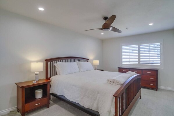 Une chambre à coucher comprenant un lit, des tables de chevet, un ventilateur de plafond, une fenêtre avec des stores et un sol recouvert de moquette.