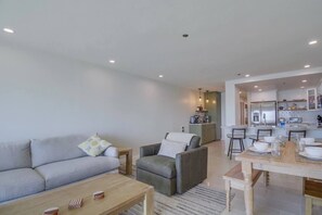 Un salon moderne avec un canapé, un fauteuil et une table basse. On aperçoit une cuisine à l’arrière-plan, avec un coin repas.