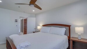 Une chambre d’hôtel avec un lit simple, une tête de lit en bois, deux lampes de chevet, un ventilateur de plafond et une porte donnant sur une autre pièce.