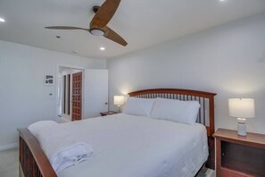 A hotel room with a single bed, a wooden headboard, two bedside lamps, a ceiling fan, and a door leading to another room.