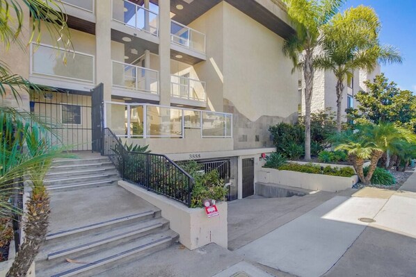 A multi-story building with a central staircase, outdoor seating area, and a small garden with palm trees.