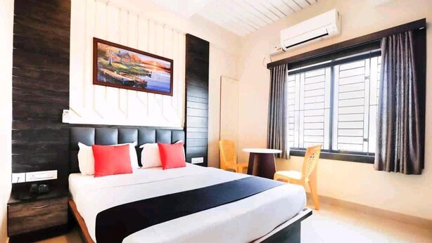 A hotel room with a large bed, a painting on the wall, a window with curtains, and a small table with a chair. - HOTEL PLAZA AVENUE  (Mangaluru)