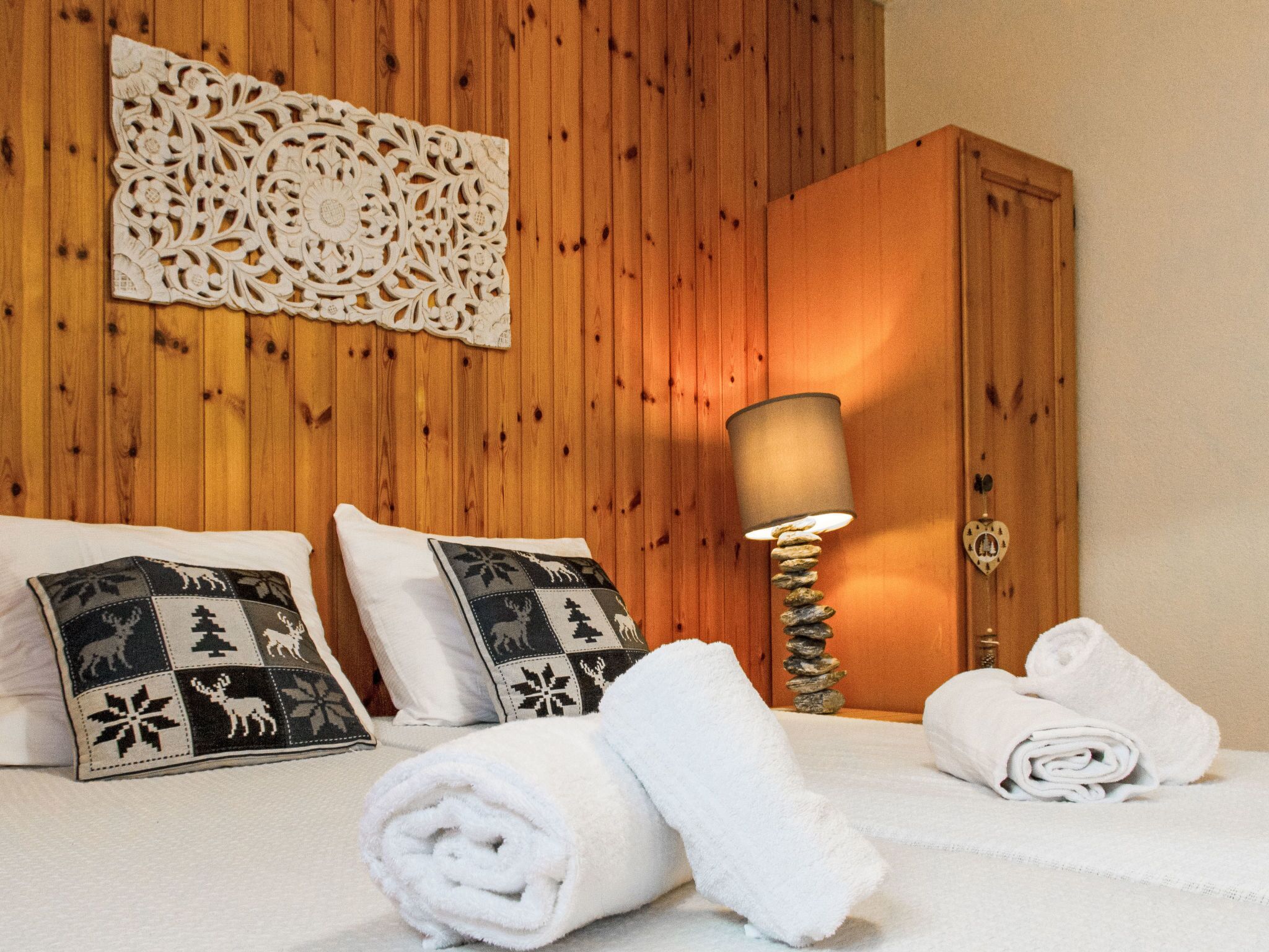 Un lit bien fait, agrémenté de deux coussins décoratifs, une pièce décorative fixée au mur, une armoire en bois et un lampadaire avec un pied en pierre.