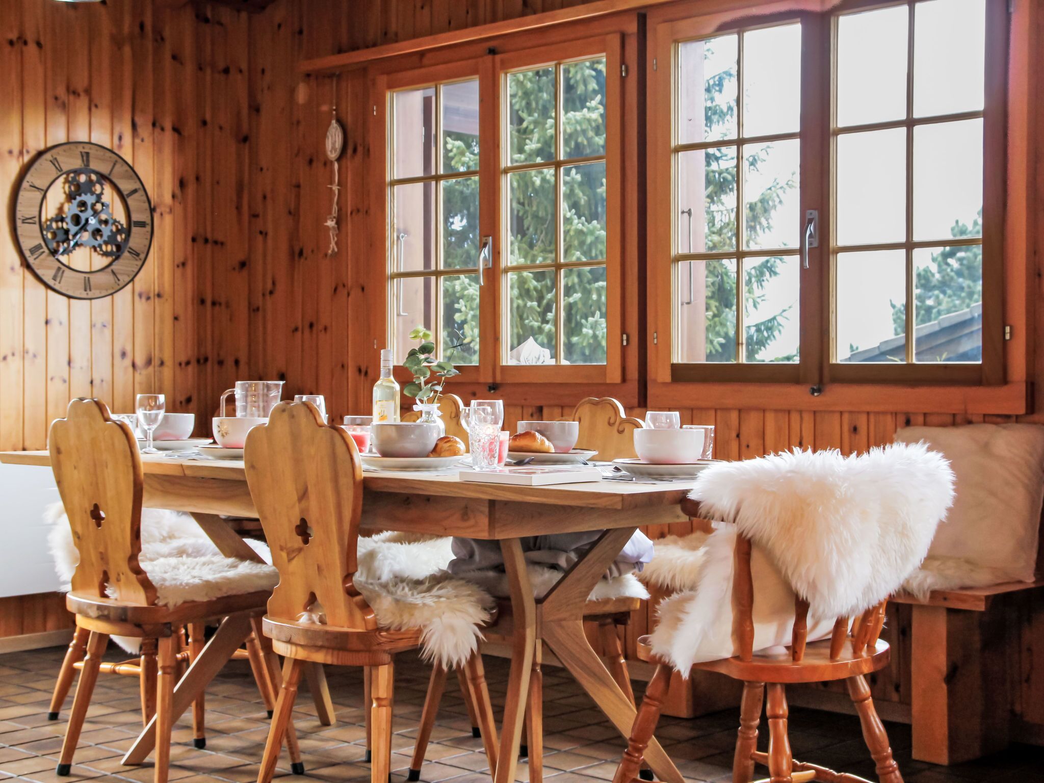 Une salle à manger en bois, avec une grande table dressée pour le petit-déjeuner, des chaises recouvertes de fausse fourrure blanche, une horloge au mur et de grandes fenêtres donnant sur la forêt.