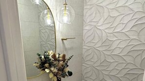 Ein Badezimmer mit einem weißen Waschbecken, einer Toilette und einer Wand mit Blättermotiv. Auf der Ablagefläche des Waschbeckens steht eine Vase mit Blumen.