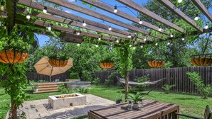 Een houten pergola met lichtjes, een vuurkorf en een houten tafel met stoelen.