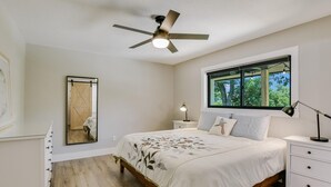 Een slaapkamer met een bed, nachtkastje, lamp, plafondventilator, raam met uitzicht en een spiegel.