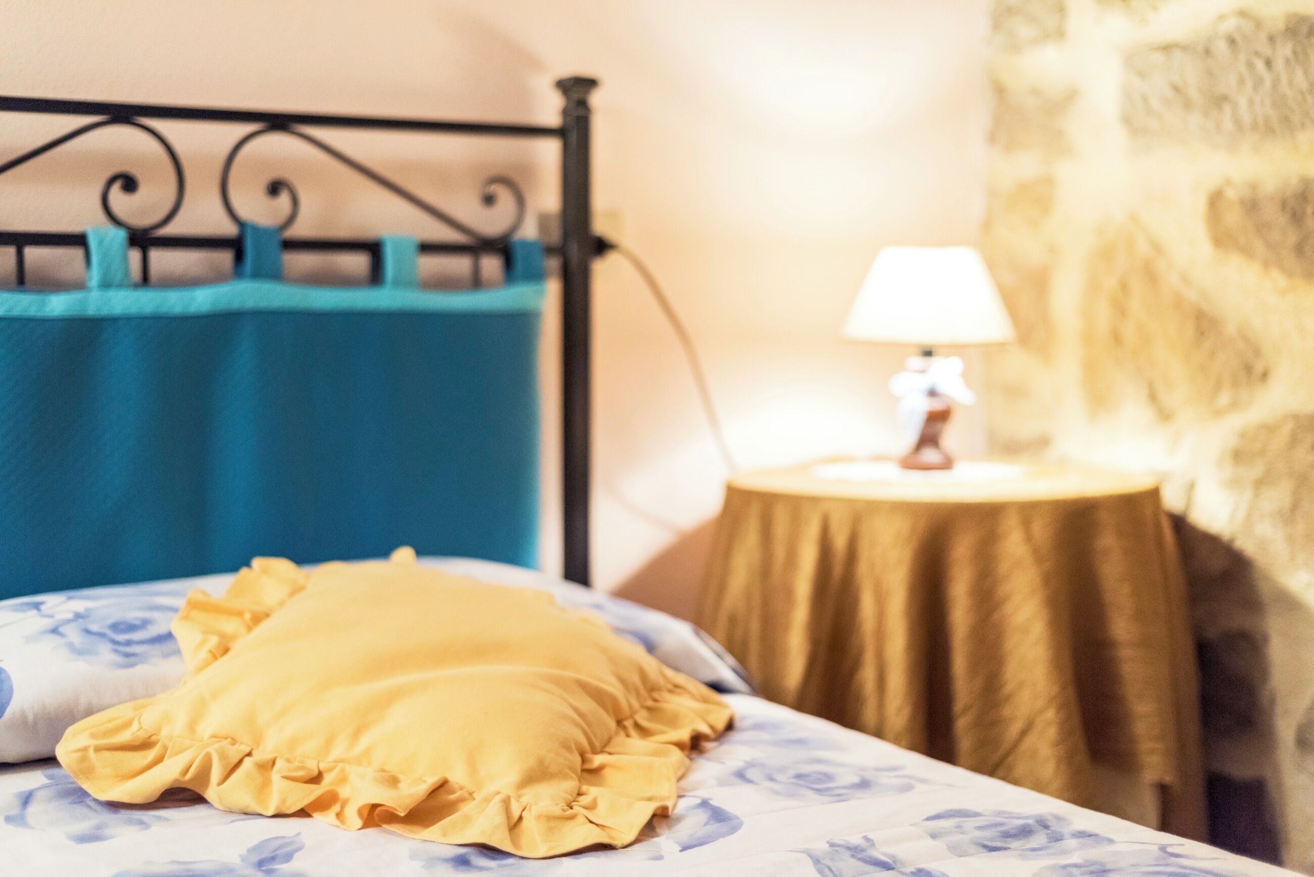 A bed with a floral bedspread, a yellow pillow, and a teal curtain. There is a lamp on a small table beside the bed.