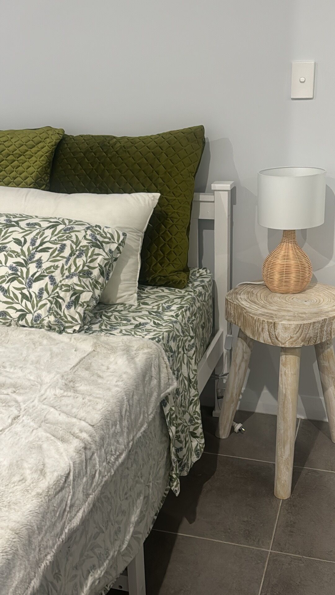 A bed with a floral bedspread, a green quilted pillow, and a white pillow. There is a small round wooden table with a woven lamp beside the bed.