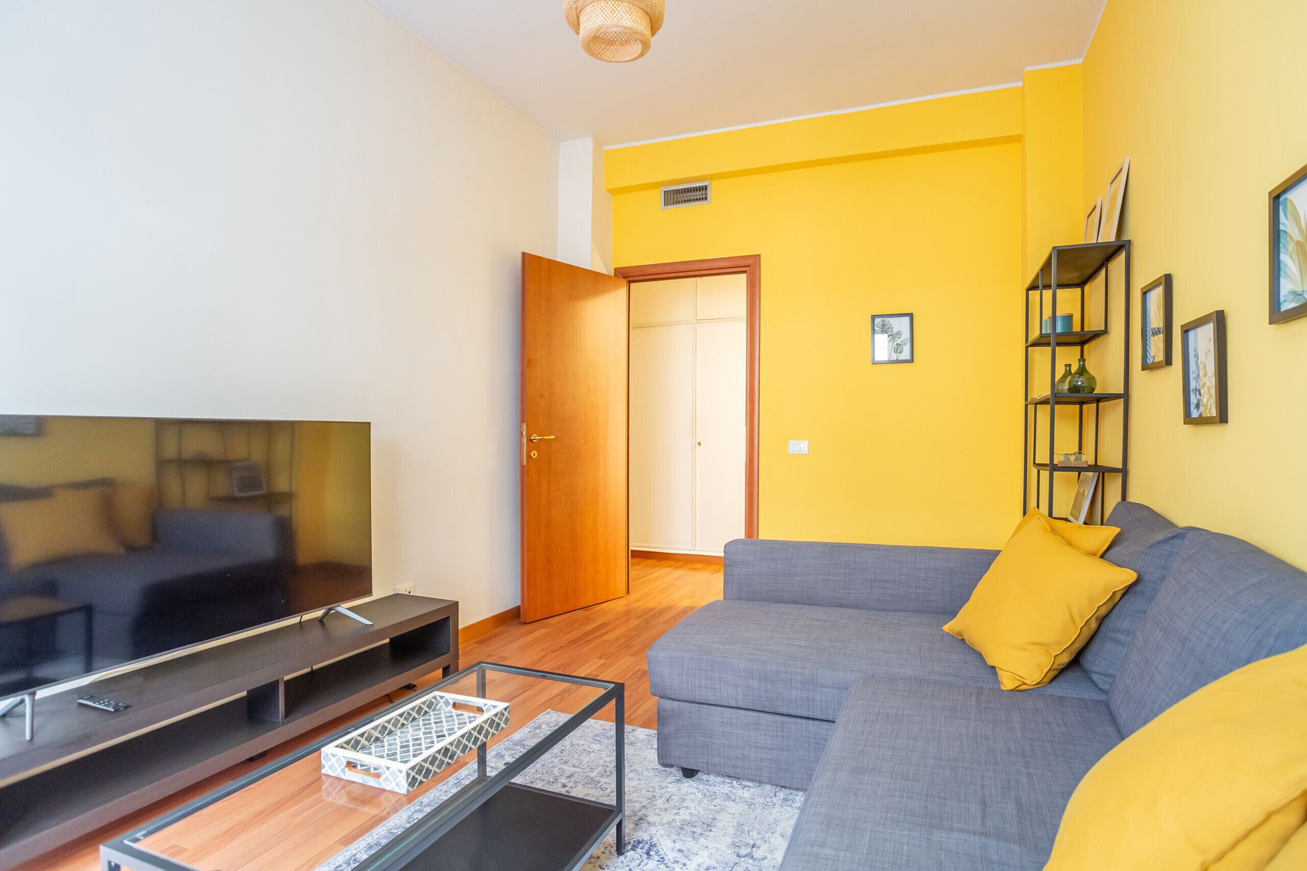 A living room with a grey sofa, yellow cushions, a TV on a stand, and a bookshelf.