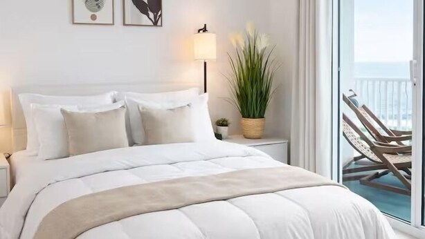 Una cama bien hecha con edredón blanco y beige, una lámpara de noche, una planta en maceta y vistas al balcón con sillas.