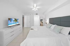 A modern hotel room with a large bed, a TV on a white dresser, a ceiling fan, and a door leading to another room.