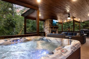 A hot tub with a stone fireplace, outdoor seating, and a TV screen.