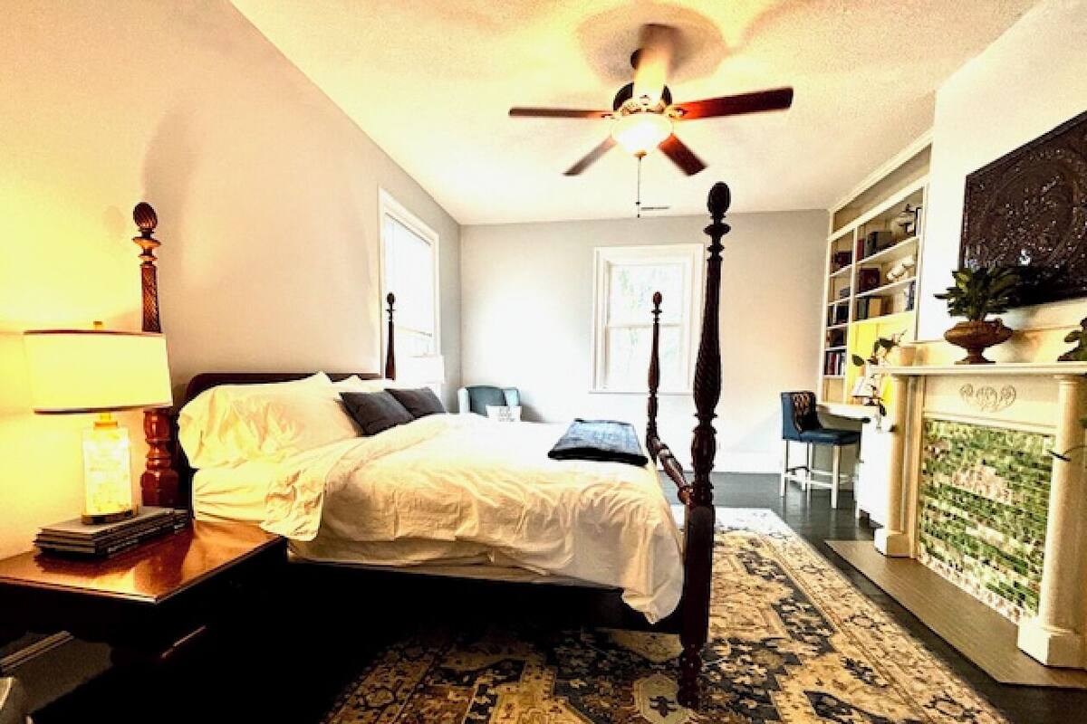 A bedroom with a four-poster bed, a ceiling fan, a bedside table with a lamp, a bookshelf, and a fireplace.