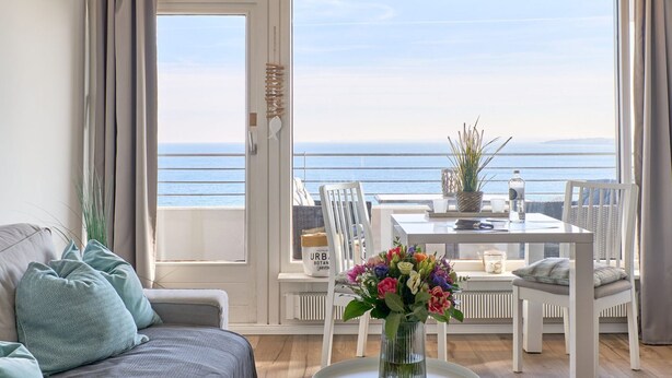 A balcony with a view of the sea, a dining table with chairs, and a sofa with cushions.