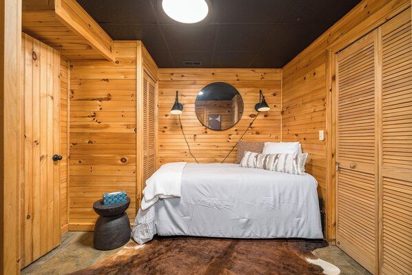 A wooden-paneled room with a bed, a round mirror, two wall-mounted lamps, and a small stool.