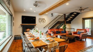 Uma sala de jantar com uma mesa de madeira comprida posta para uma refeição, uma lareira e uma escada que leva a um andar superior.