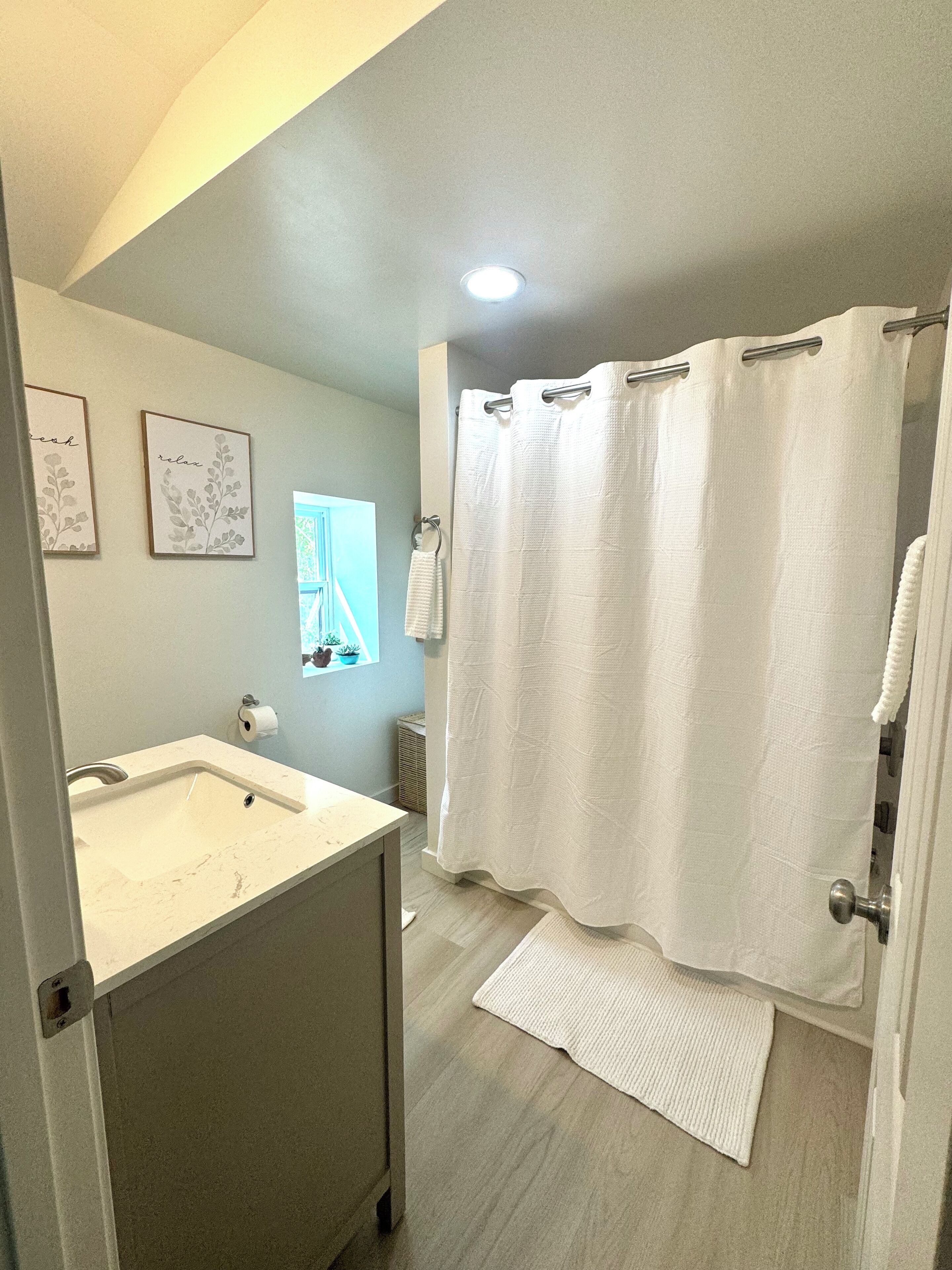 A bathroom with a sink, a shower curtain, a towel rack, and a small window with a plant.
