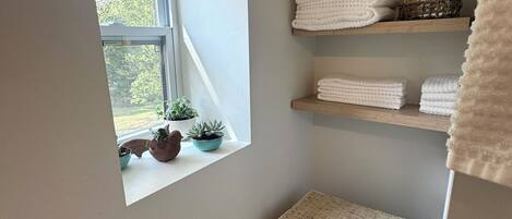 Un baño con ventana, repisas con toallas, una cesta de mimbre y una planta en maceta.