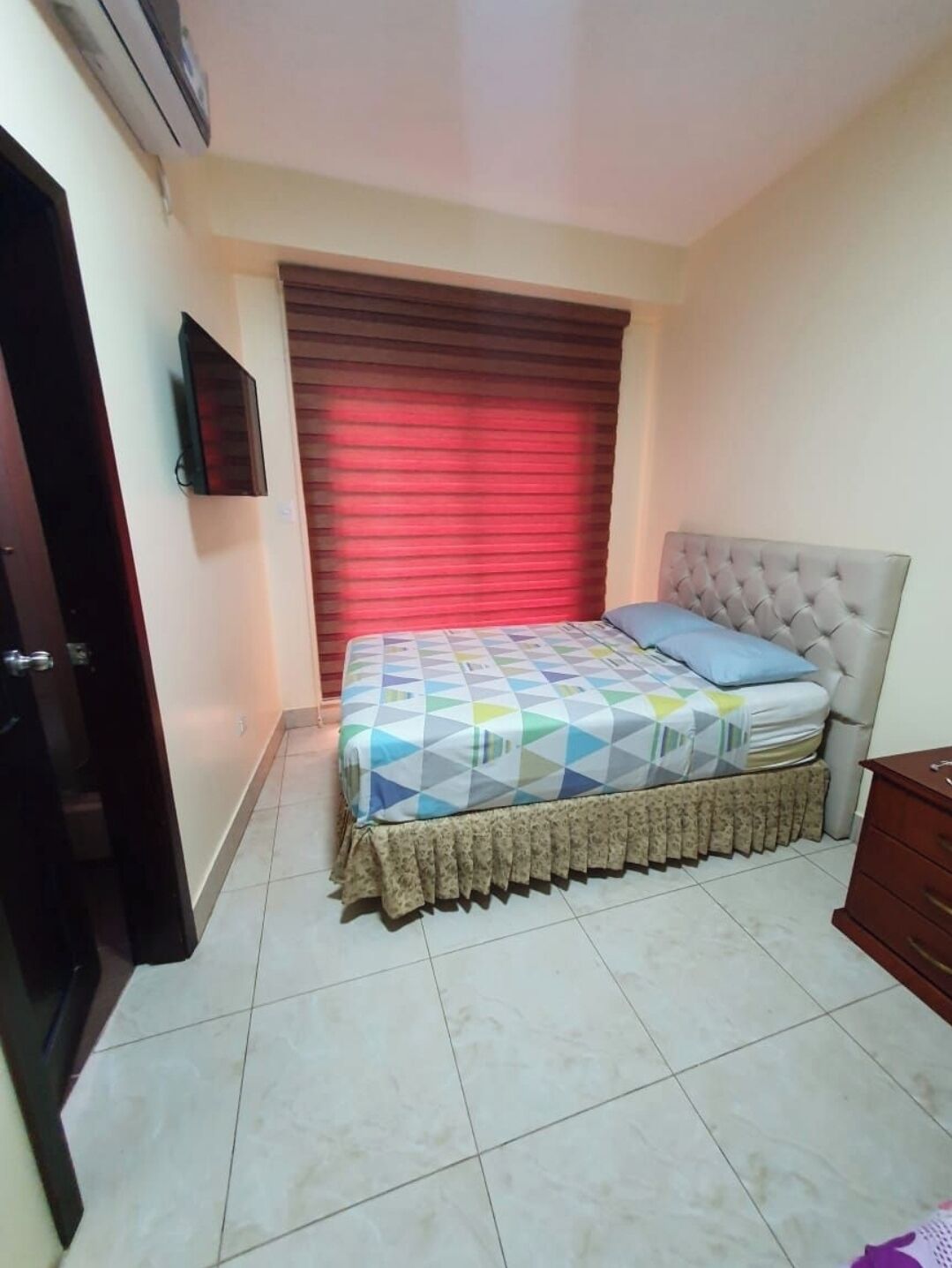 Un dormitorio con una cama, un televisor montado en la pared y una ventana con persianas rojas.