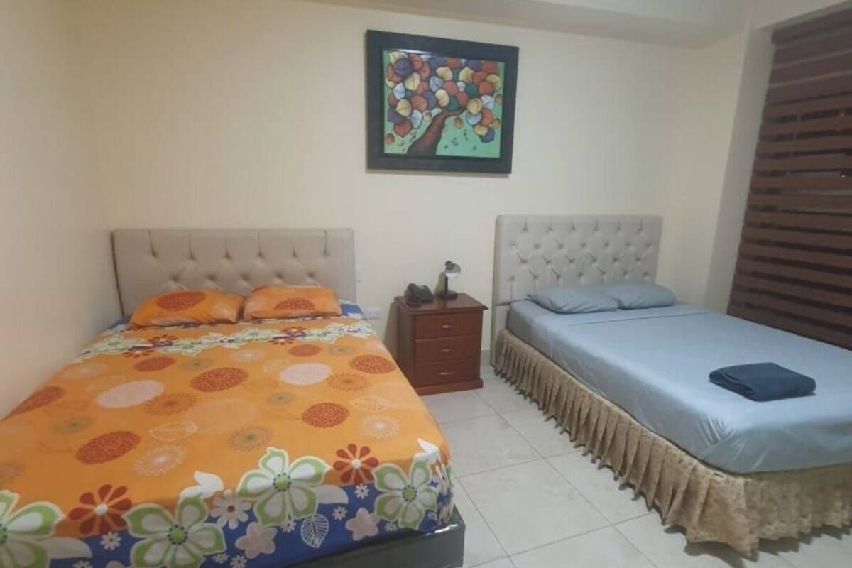 Un dormitorio con dos camas, una con cubrecama naranja con estampado floral y la otra con cubrecama azul. Hay una mesita de noche de madera entre las camas, y un cuadro enmarcado cuelga de la pared sobre ellas.