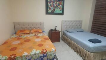 Quarto com duas camas, uma com colcha florida laranja e outra com colcha azul. Há uma pequena mesa de cabeceira de madeira entre as camas, e um quadro emoldurado está pendurado na parede acima delas.