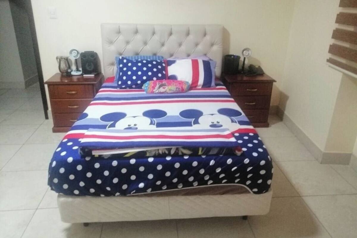 Un dormitorio con una cama con edredón de temática de Mickey Mouse, dos mesitas de noche con altavoces y una obra de arte colgada en la pared.