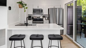 Una cocina moderna con gabinetes blancos, electrodomésticos de acero inoxidable, una isla central con taburetes y un ventanal corredizo a un patio.