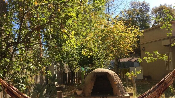 Een hangmat in een tuin met een stenen bakoven en een houten hek op de achtergrond.
