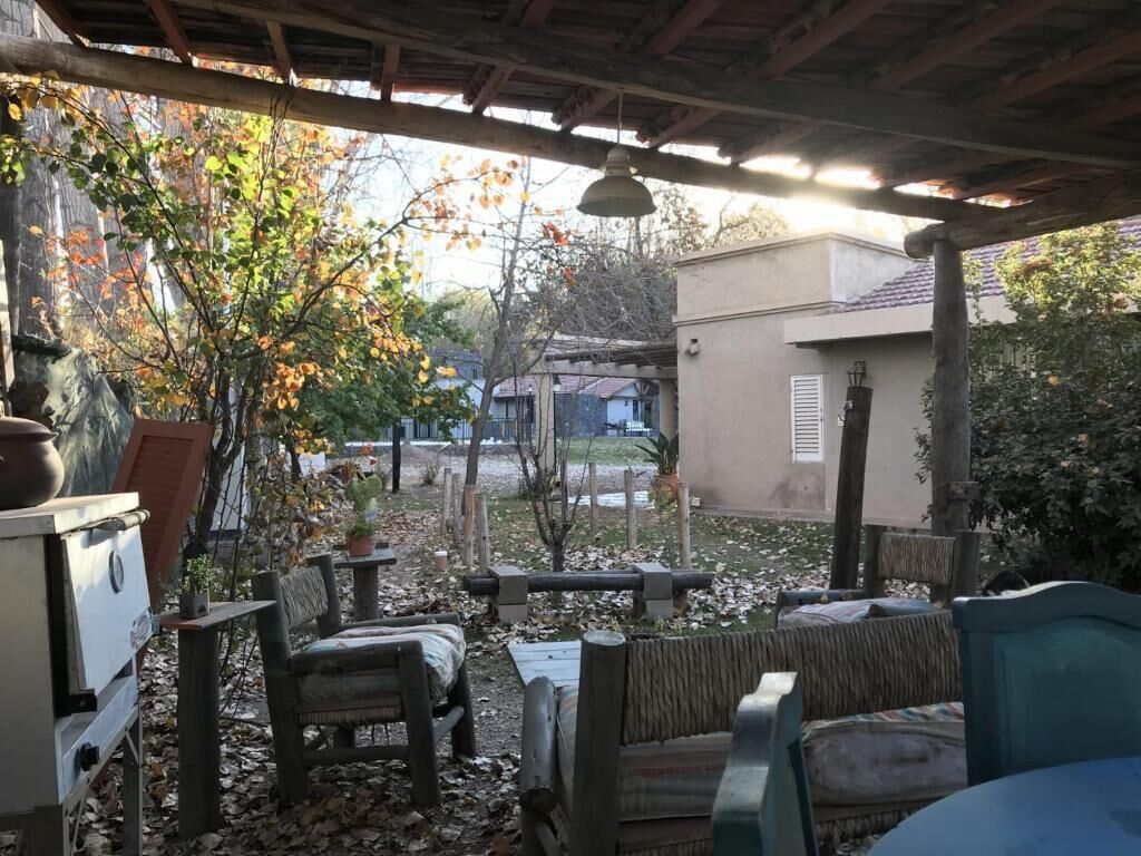 A covered outdoor seating area with wooden furniture and a table, surrounded by trees and a building.