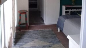 Een kleine, eenvoudige kamer met een bed, een krukje, een schilderij en een deur naar een andere kamer.