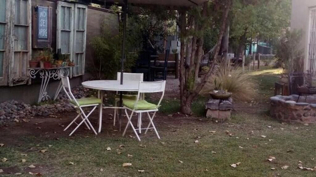 Un patio con una mesa redonda y dos sillas, rodeado por un jardín con árboles y una jardinera de piedra.