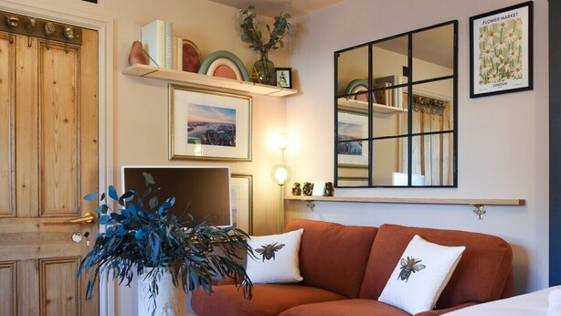 Una habitación acogedora con una puerta de madera, un sofá marrón con cojines blancos, una mesita de noche blanca con una planta y un espejo grande en la pared.