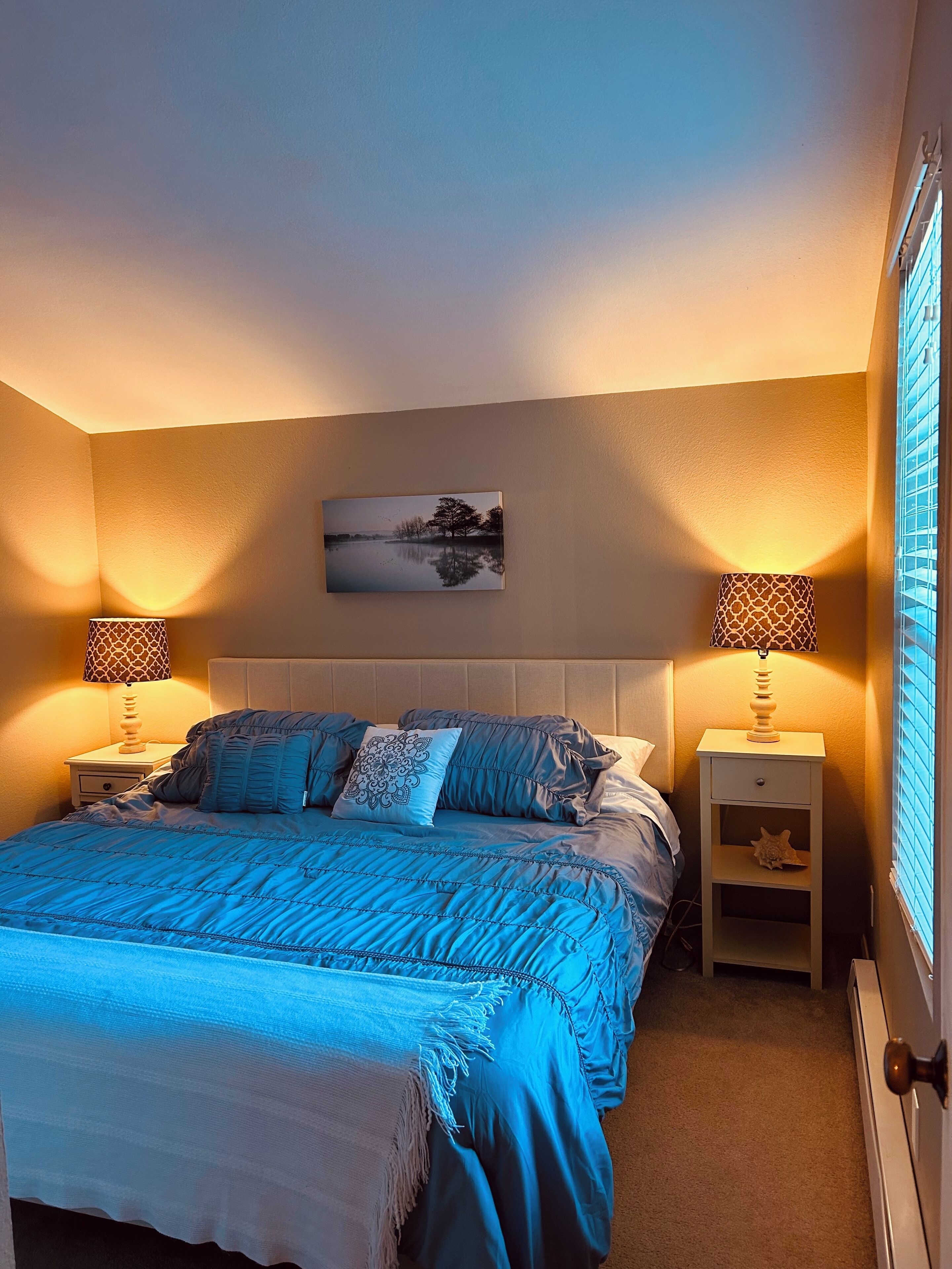 Une chambre avec un grand lit, deux tables de chevet avec des lampes, une œuvre d’art accrochée au mur et une fenêtre avec des stores.