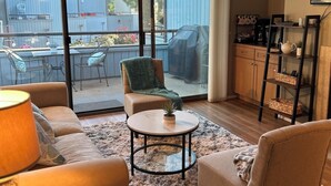 A spacious living area with a large sliding glass door leading to an outdoor patio. There is a comfortable seating area with a sofa, armchairs, and a coffee table. A television is mounted on the wall, and a kitchen area is visible in the background.